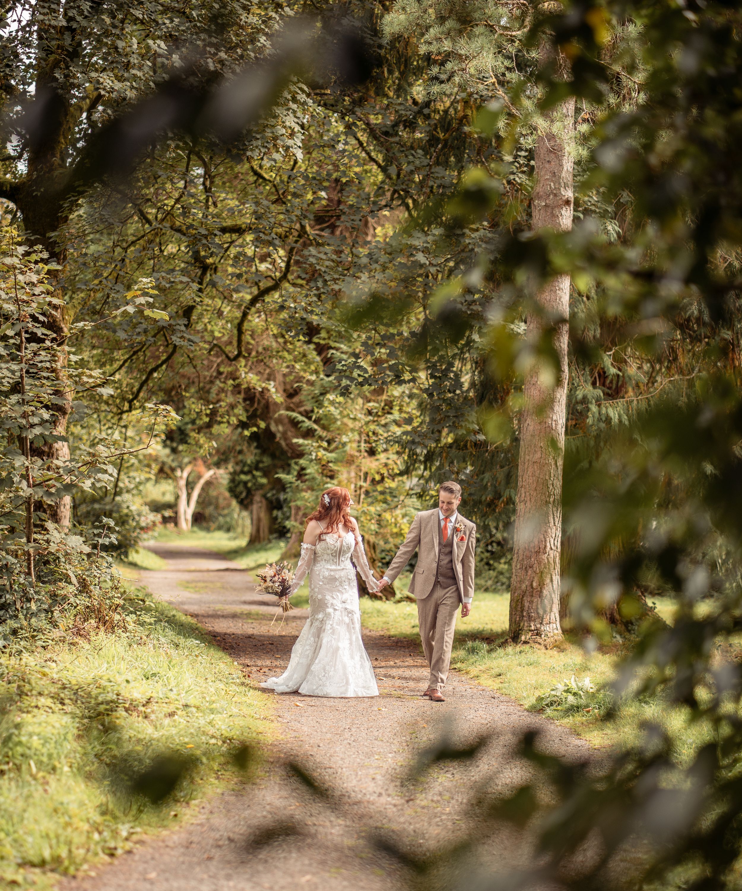 A bride and groom walk hand-in-hand along a tree-lined path in a lush green woodland setting.