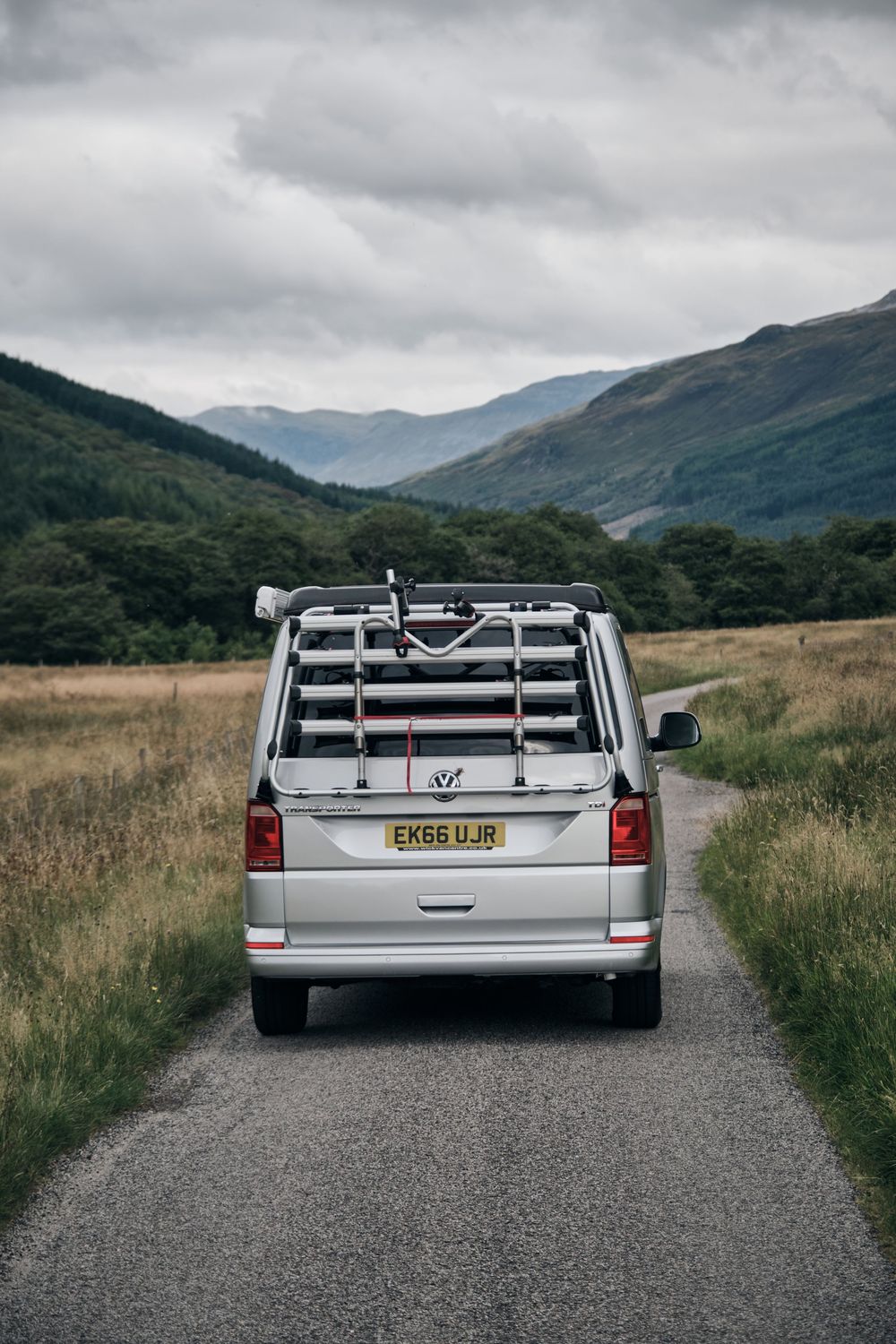 Silver VW campervan driving on a narrow road through scenic Scottish Highlands valley with mountains and cloudy sky.