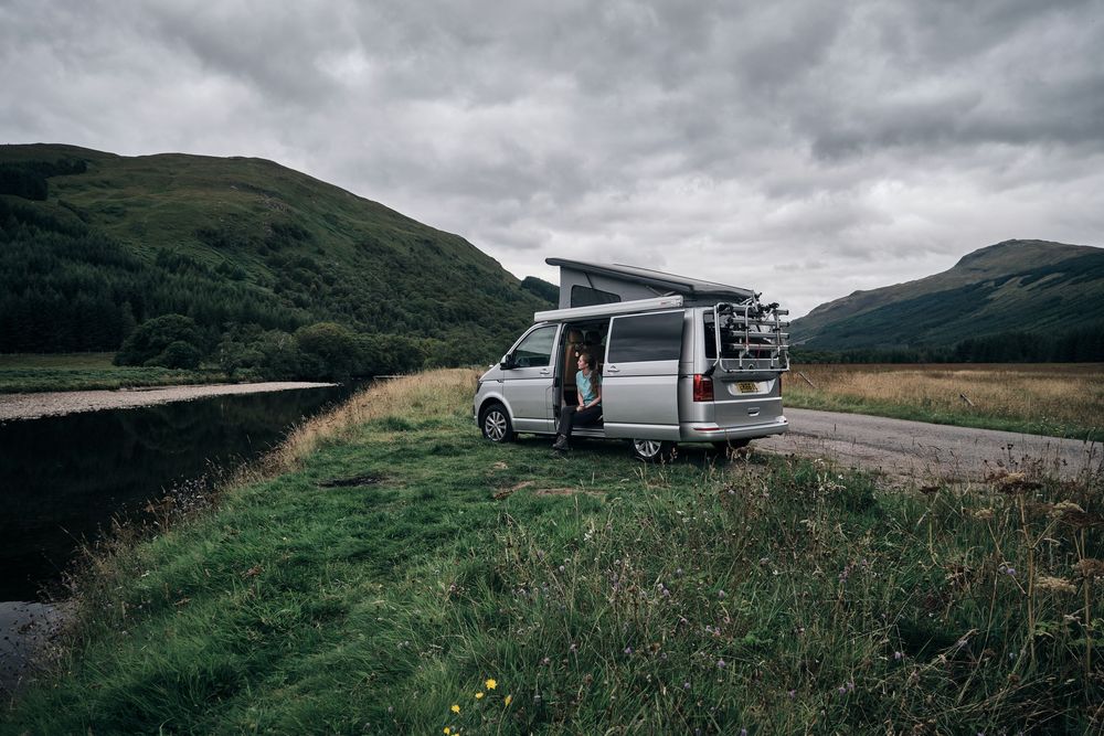 A silver VW campervan parked on a scenic highland road beside a river, with green hills and dramatic cloudy skies.
