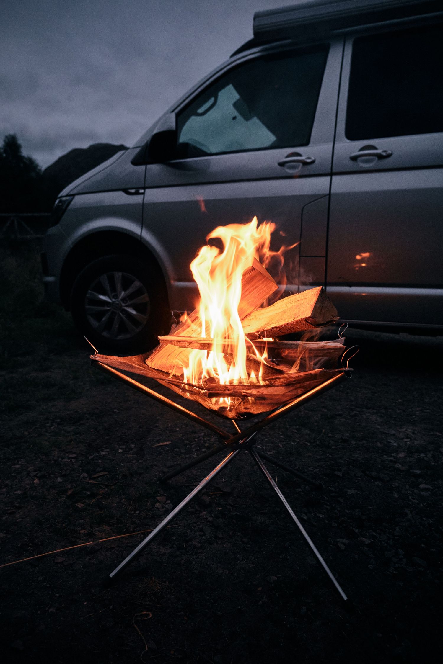 Portable campfire burning brightly on a metal stand at dusk beside a gray VW Transporter camper van in a scenic outdoor setting.