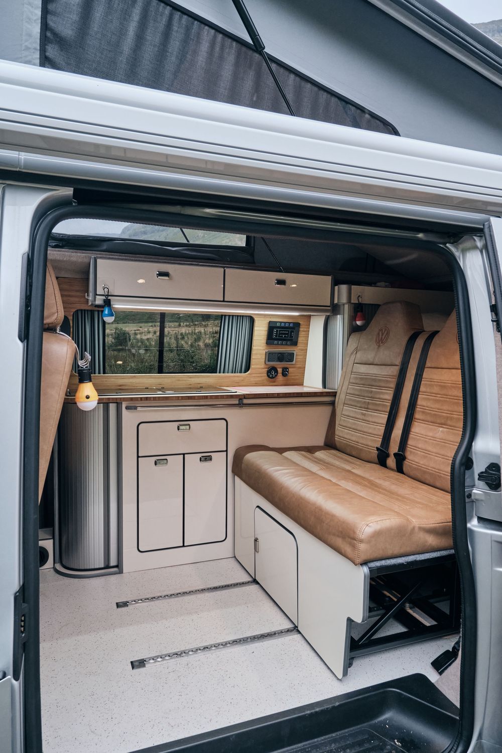 Interior view of a camper van conversion with leather bench seat, wooden cabinetry, compact kitchen, and pop-top roof.