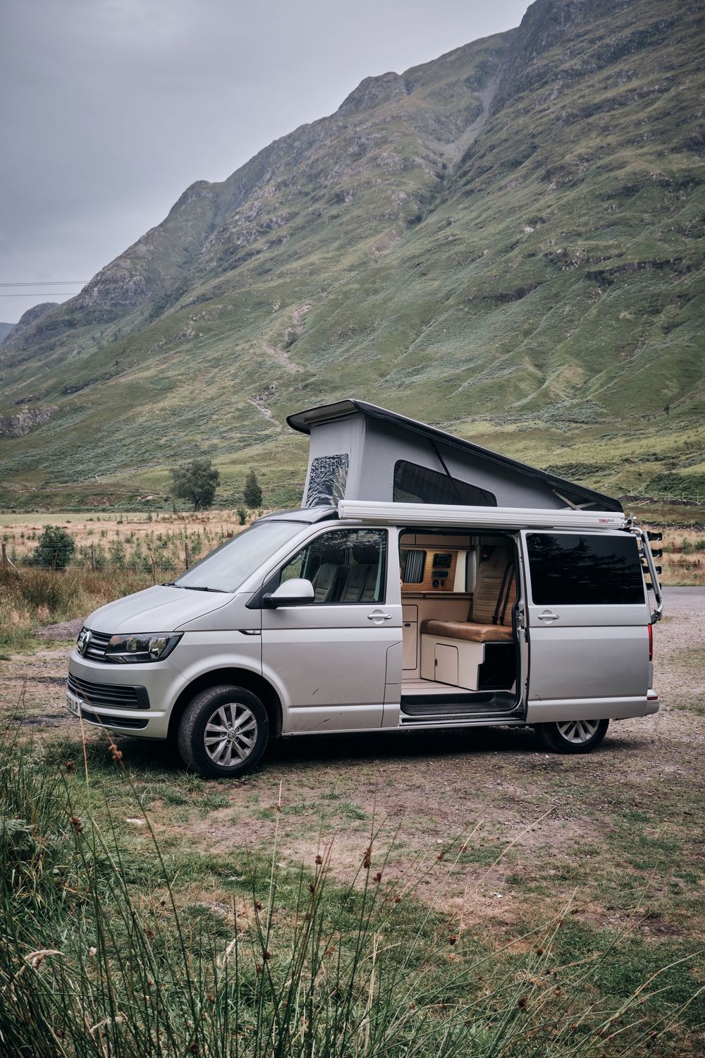 Silver VW California campervan with pop-up roof open parked in Scottish Highlands valley with dramatic green mountains backdrop.
