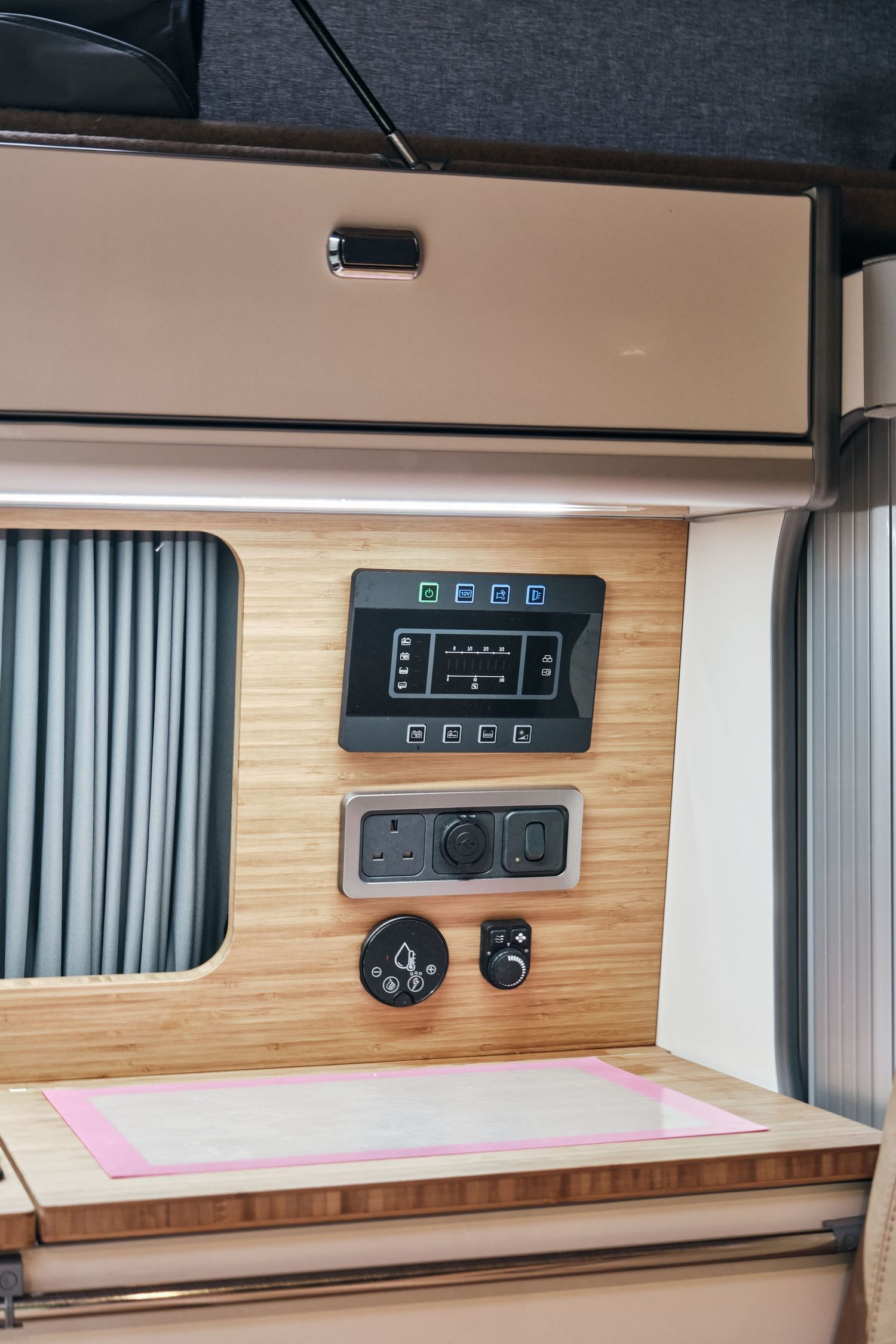Modern campervan interior with bamboo wood panels, control panel, electrical outlets, and overhead storage compartment.