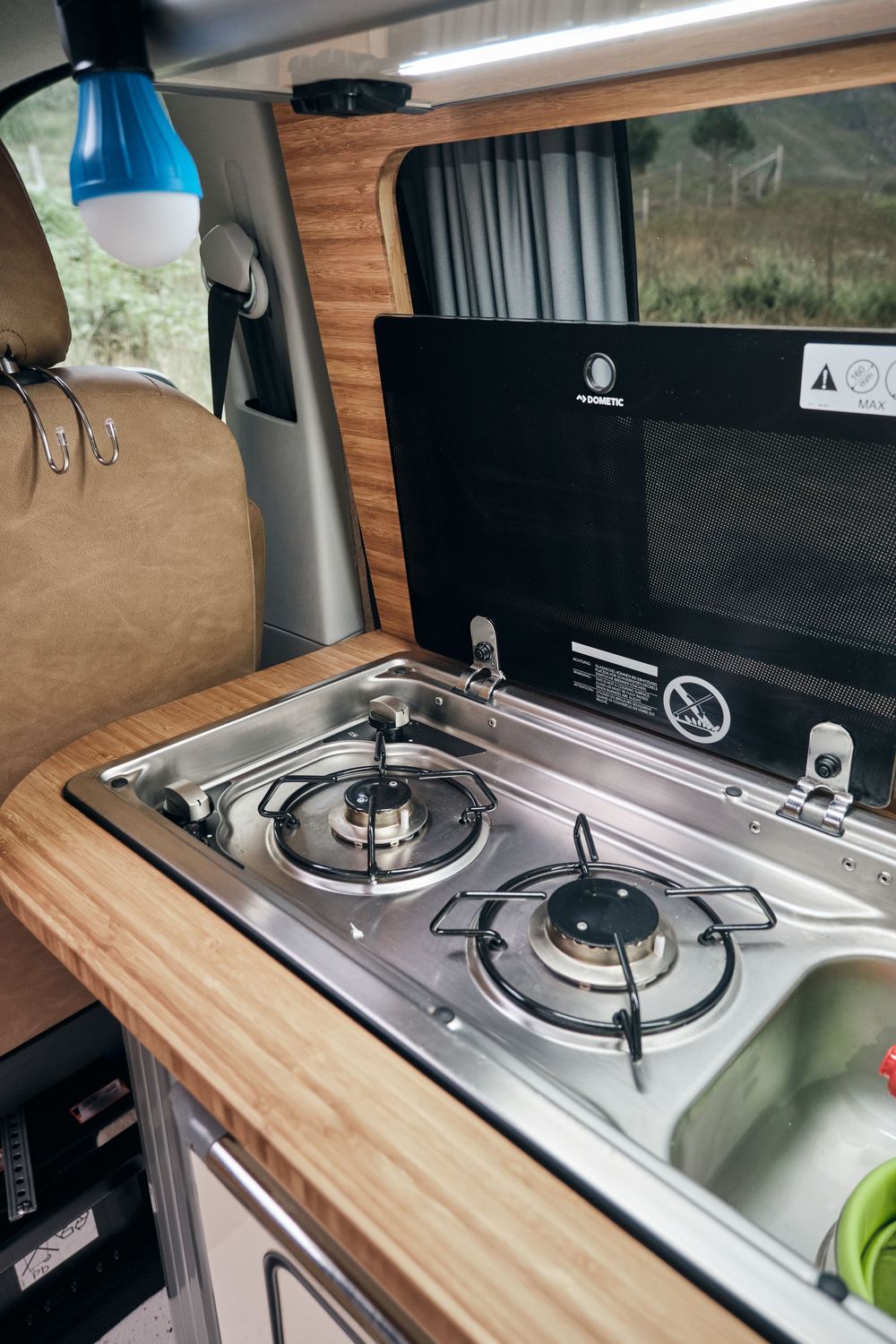 Compact two-burner gas stove with stainless steel surface and Dometic microwave in a modern camper van conversion kitchen.