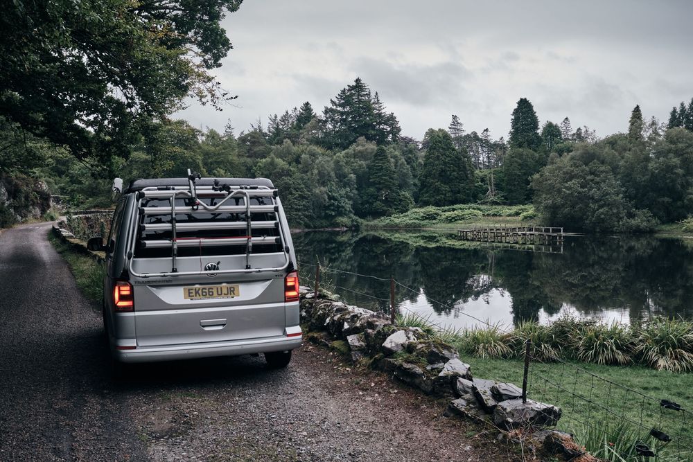 Silver campervan with bike rack parked beside a tranquil lake surrounded by lush green trees on a cloudy day.