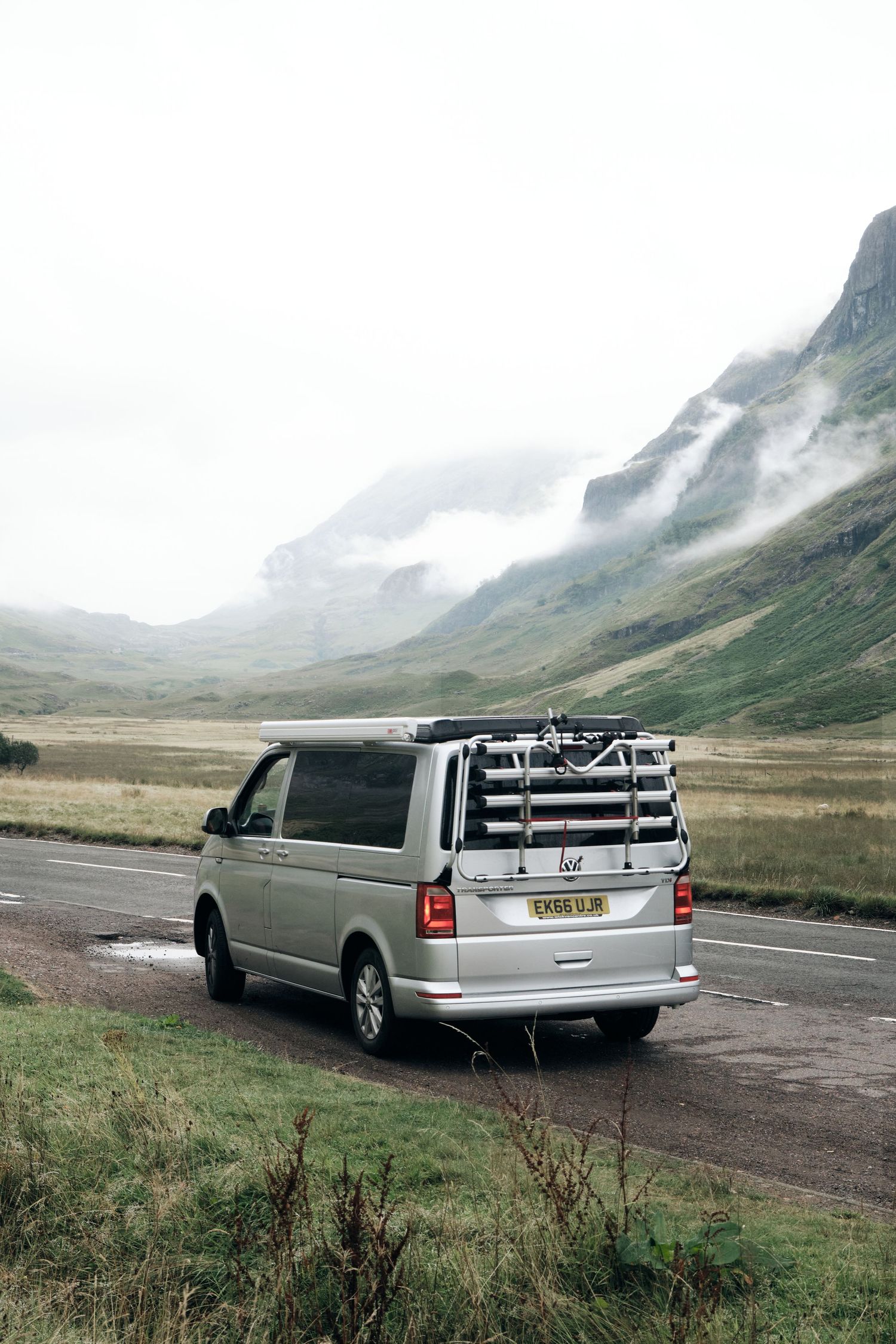 Silver VW campervan parked on a misty Scottish Highland road with dramatic mountains and green valleys in the background.