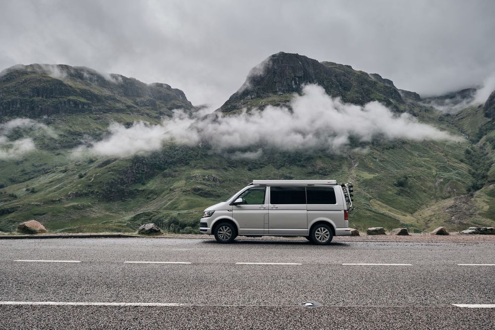 Silver VW campervan parked on mountain road with misty green peaks and low clouds in Scottish Highlands.