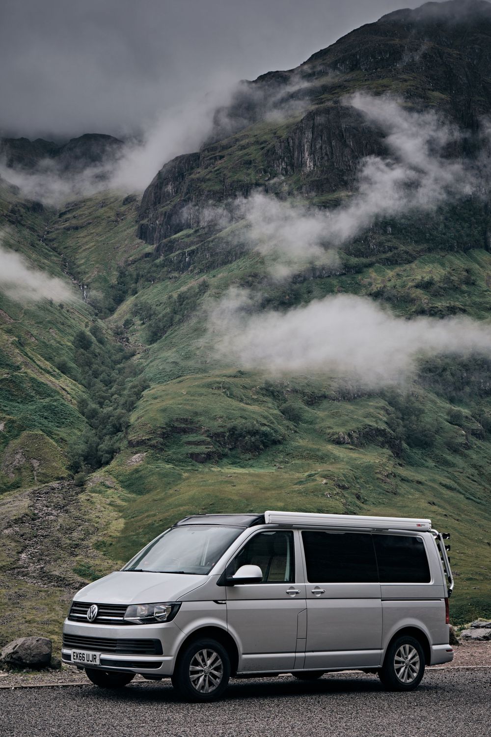 Silver VW Transporter campervan parked on a mountain road with dramatic misty green highlands in the background.