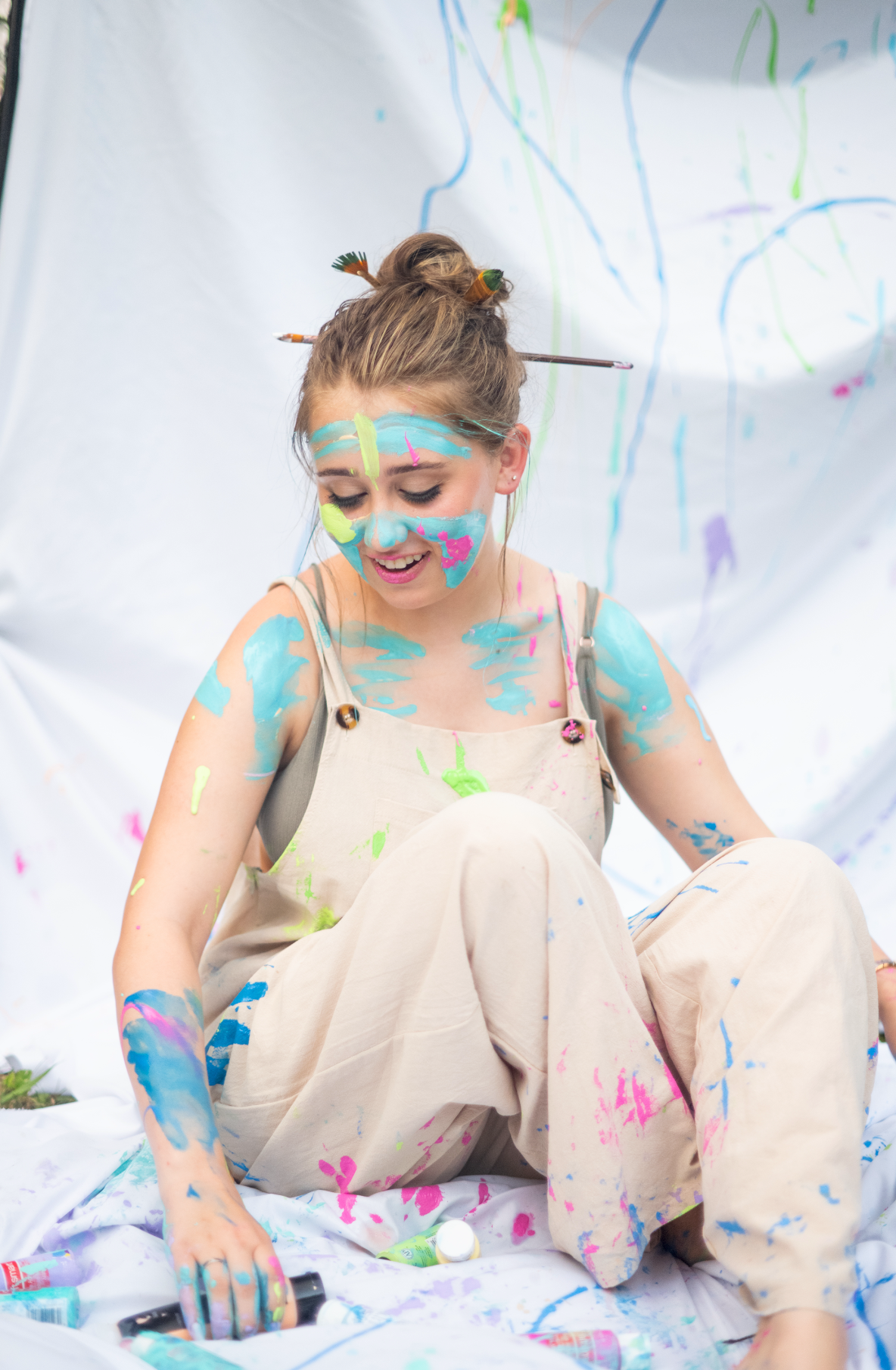 A young woman covered in colorful paint sits smiling on a white sheet, wearing beige overalls with a paintbrush in her hair.