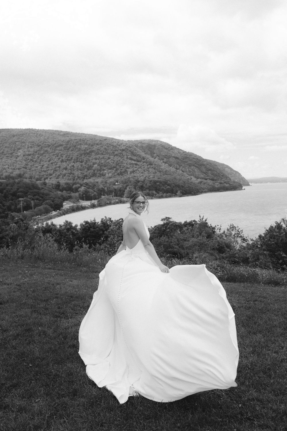 Bride in flowing white gown twirling outdoors with scenic river and mountain backdrop in black and white photo.