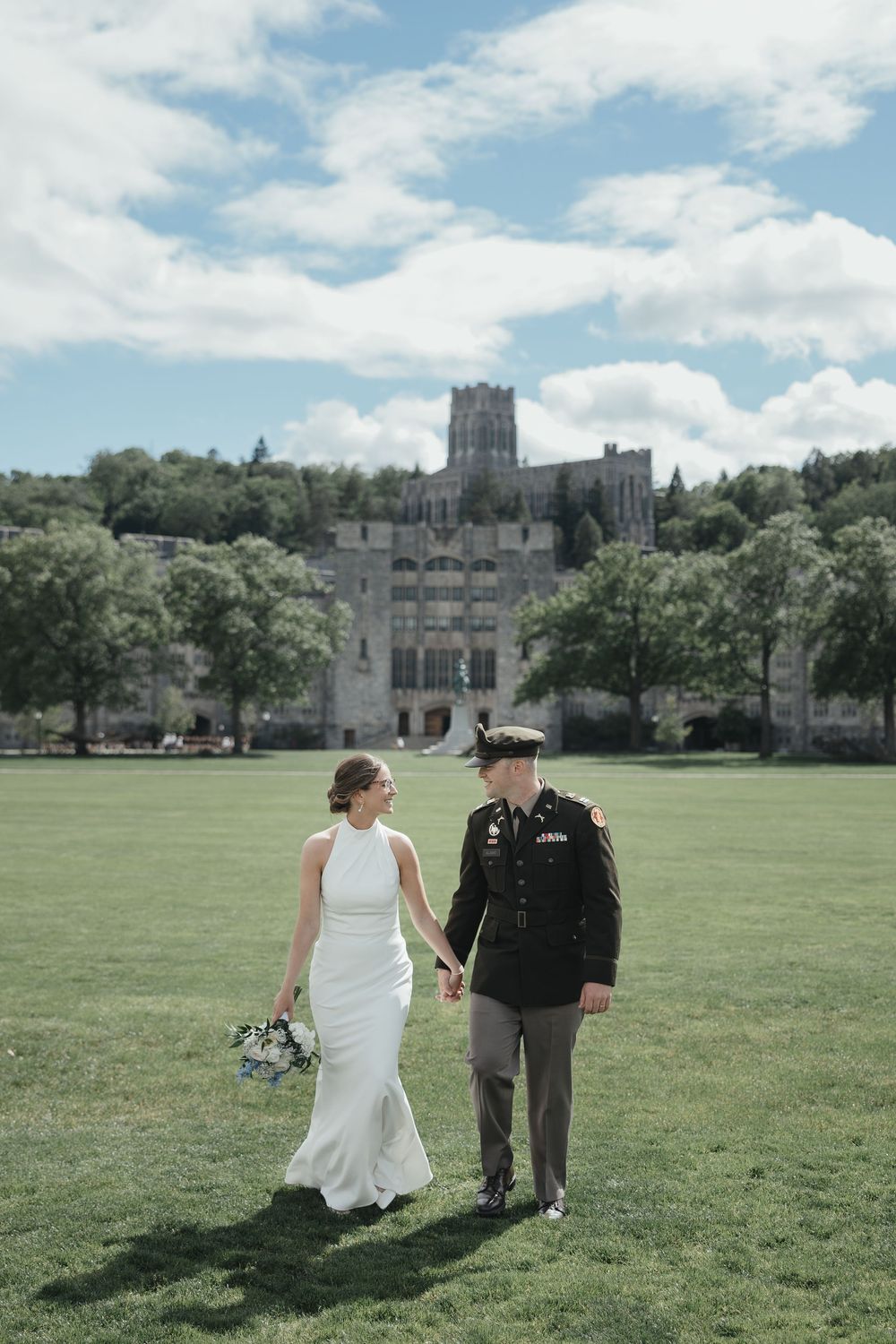 Bride and groom in military uniform holding hands on green lawn with historic West Point castle building backdrop.