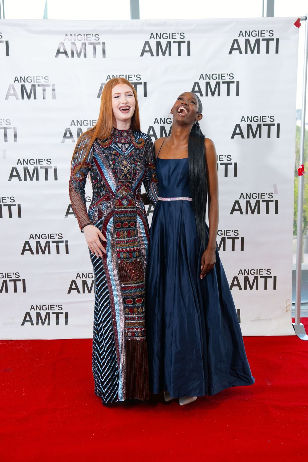 Two elegantly dressed women laughing together on a red carpet at Angie's AMTI event.