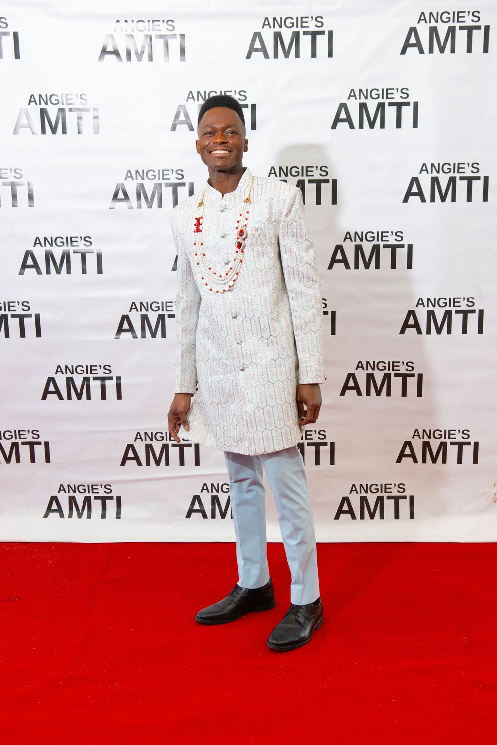 Man in white traditional African outfit with red beaded necklace poses on red carpet at Angies AMTI event.