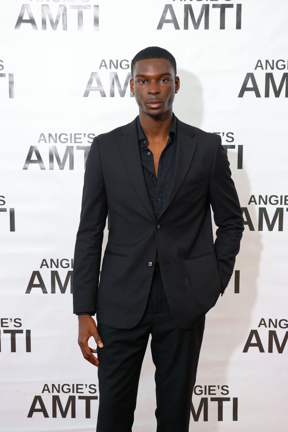 Tall man in black suit poses confidently at Angie's AMTI fashion event backdrop.