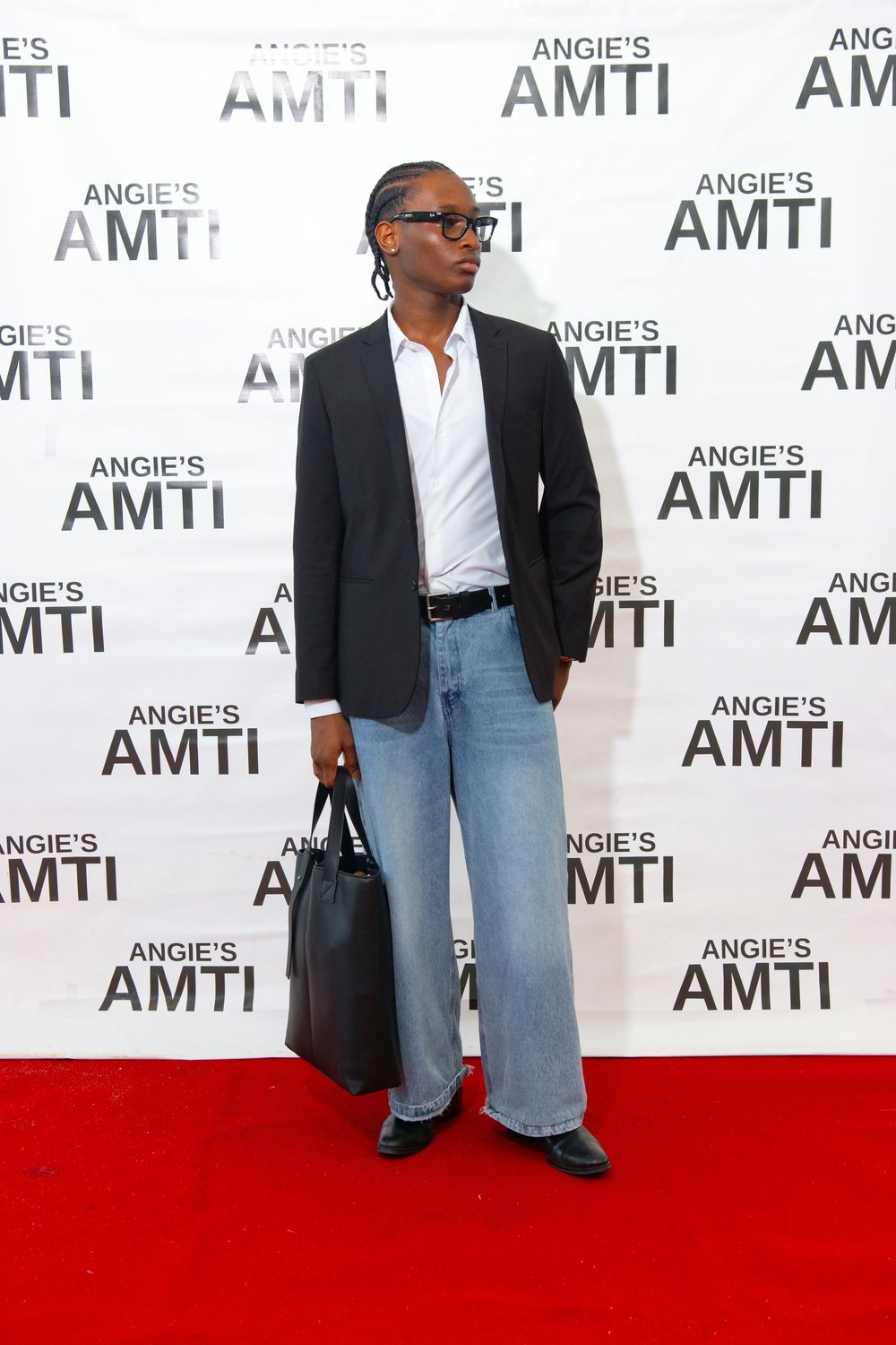 Stylish person in black blazer, white shirt, wide-leg jeans holds black bag on Angies AMTI red carpet.