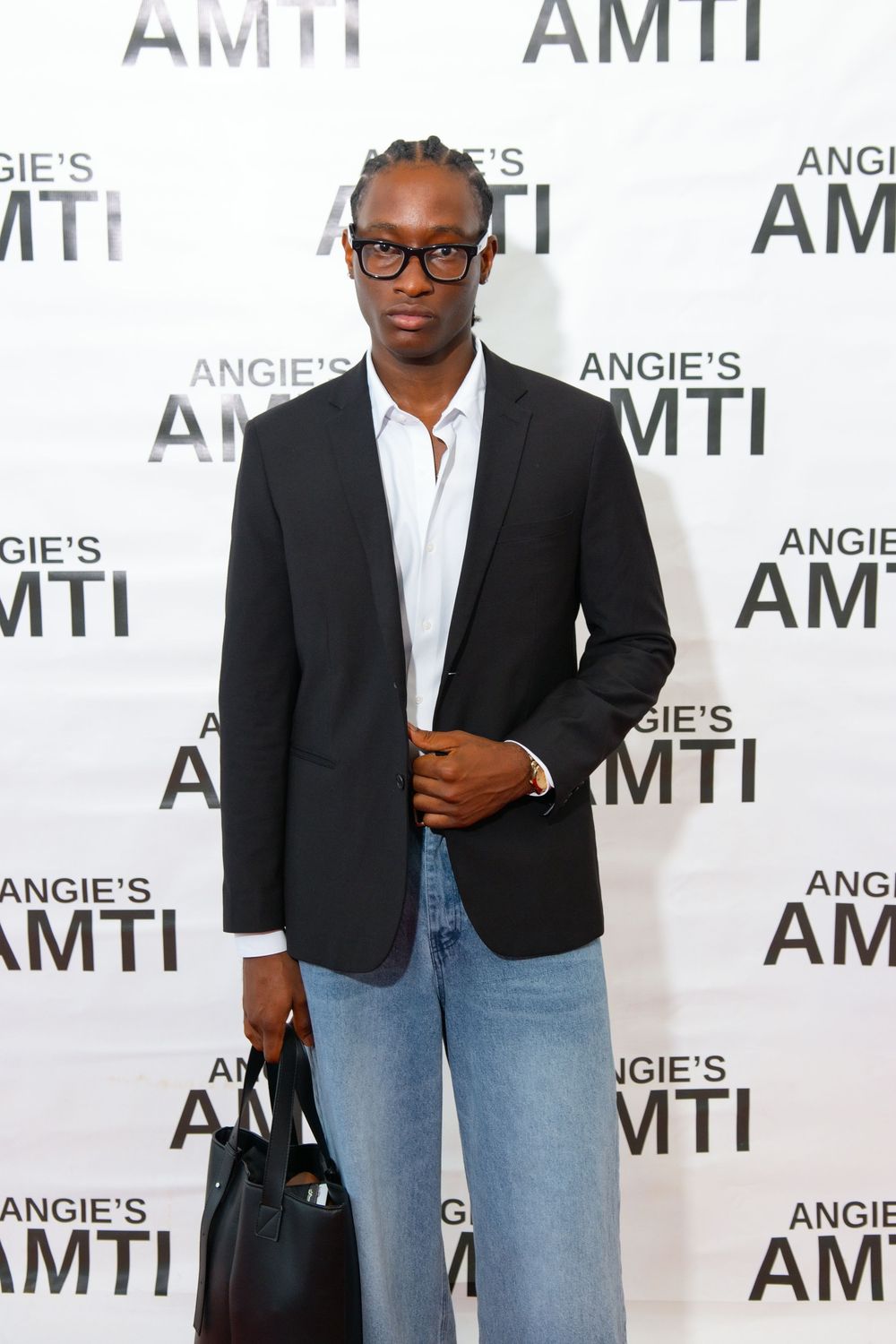 Man in black blazer, jeans, and glasses poses at Angie's AMTI event backdrop holding a black bag.