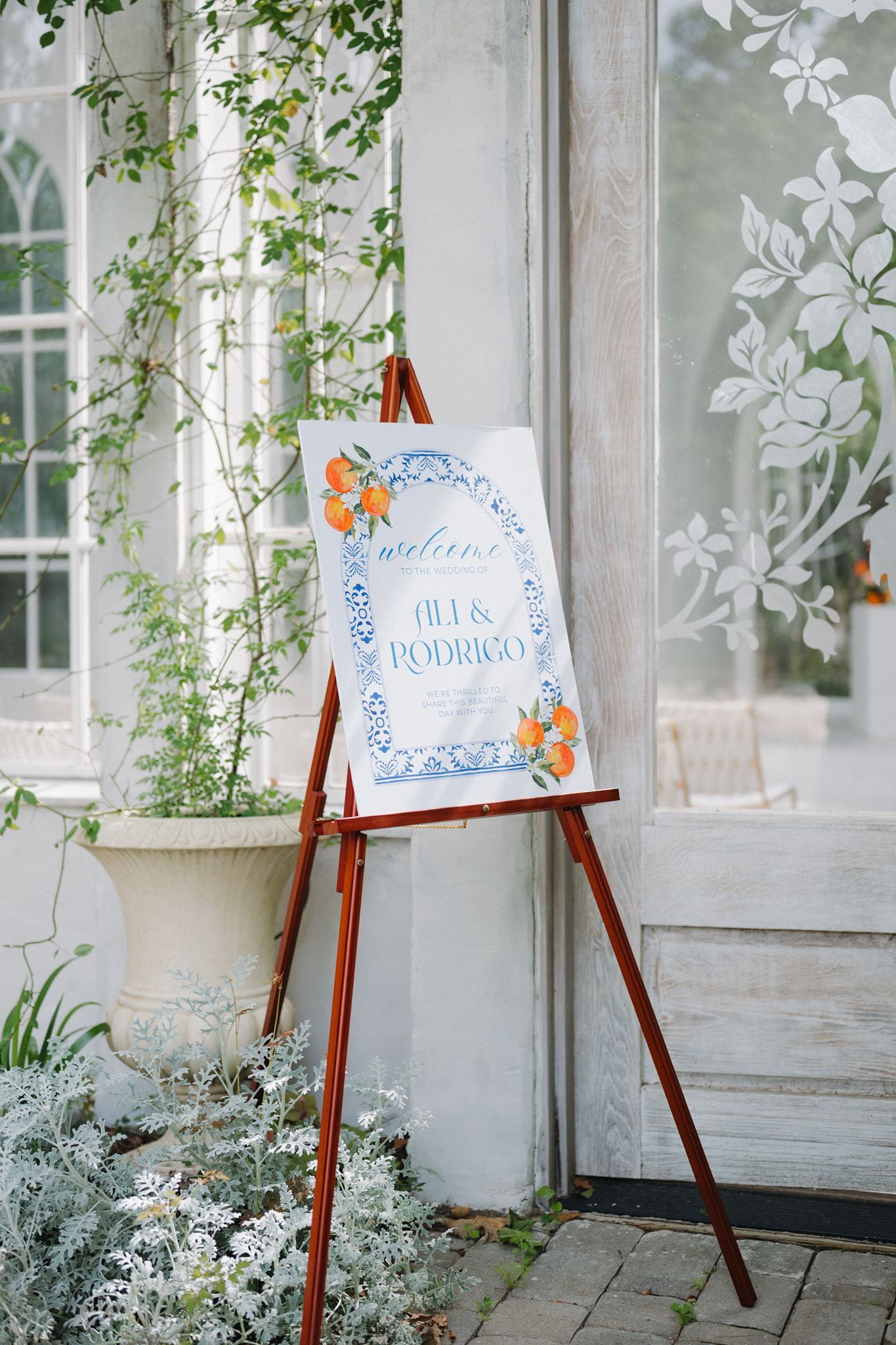 Wedding welcome sign for Ali and Rodrigo on a wooden easel, featuring blue floral border and orange accents.