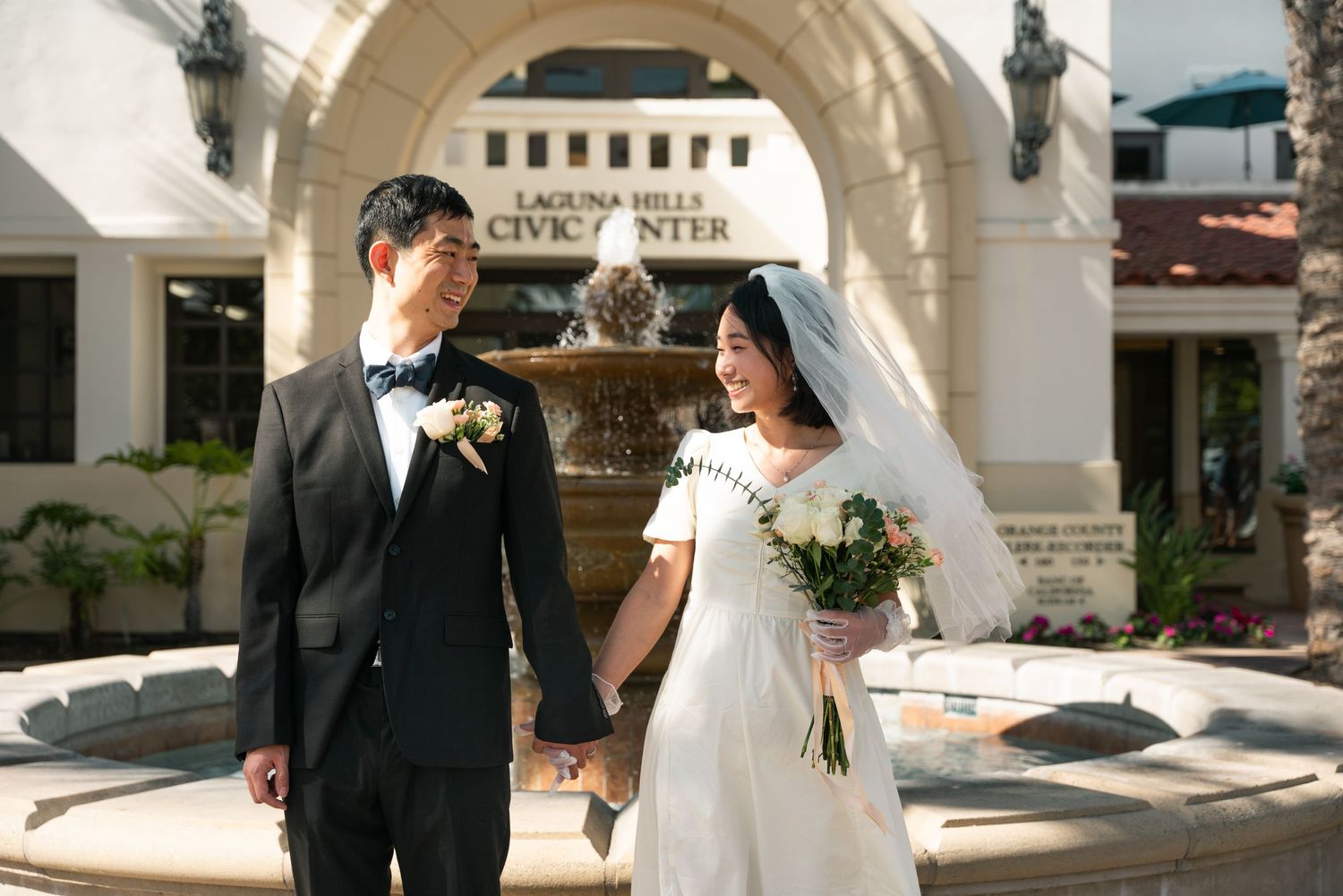 A couple in wedding attire holds hands and smiles at each other near a fountain at Laguna Hills Civic Center.