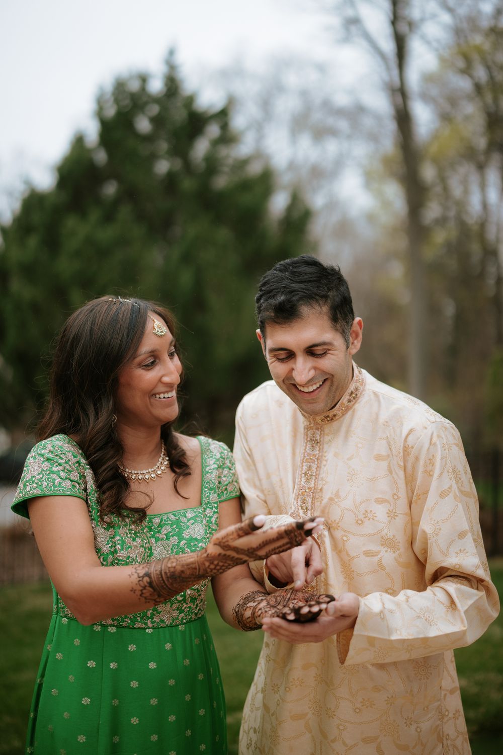 Indian couple in traditional attire smiling, bride in green salwar with henna hands, groom in cream sherwani, outdoors.
