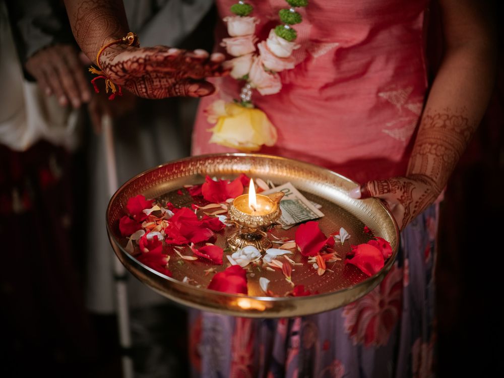 Hands with henna holding a brass plate with floating rose petals and lit diya candle at an Indian wedding ceremony.