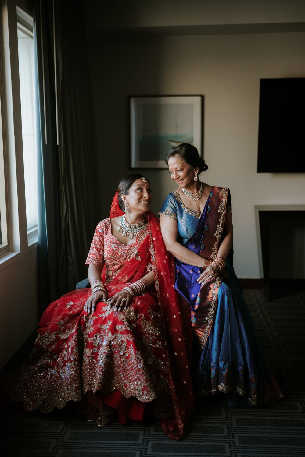 Bride in red lehenga and woman in blue saree sitting together by window in Indian wedding preparation.