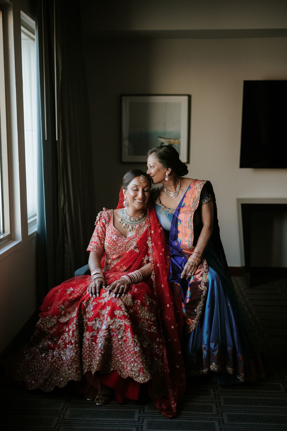 Indian bride in red lehenga sits by window with older woman in blue saree during bridal wedding preparation.
