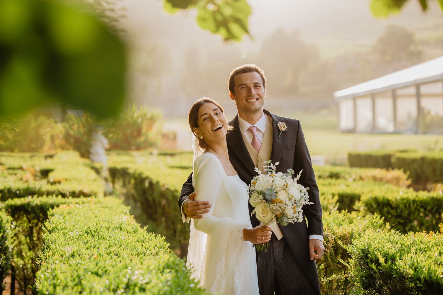 Happy bride and groom pose in a sunlit formal garden, bride holding a white bouquet in an elegant wedding dress.
