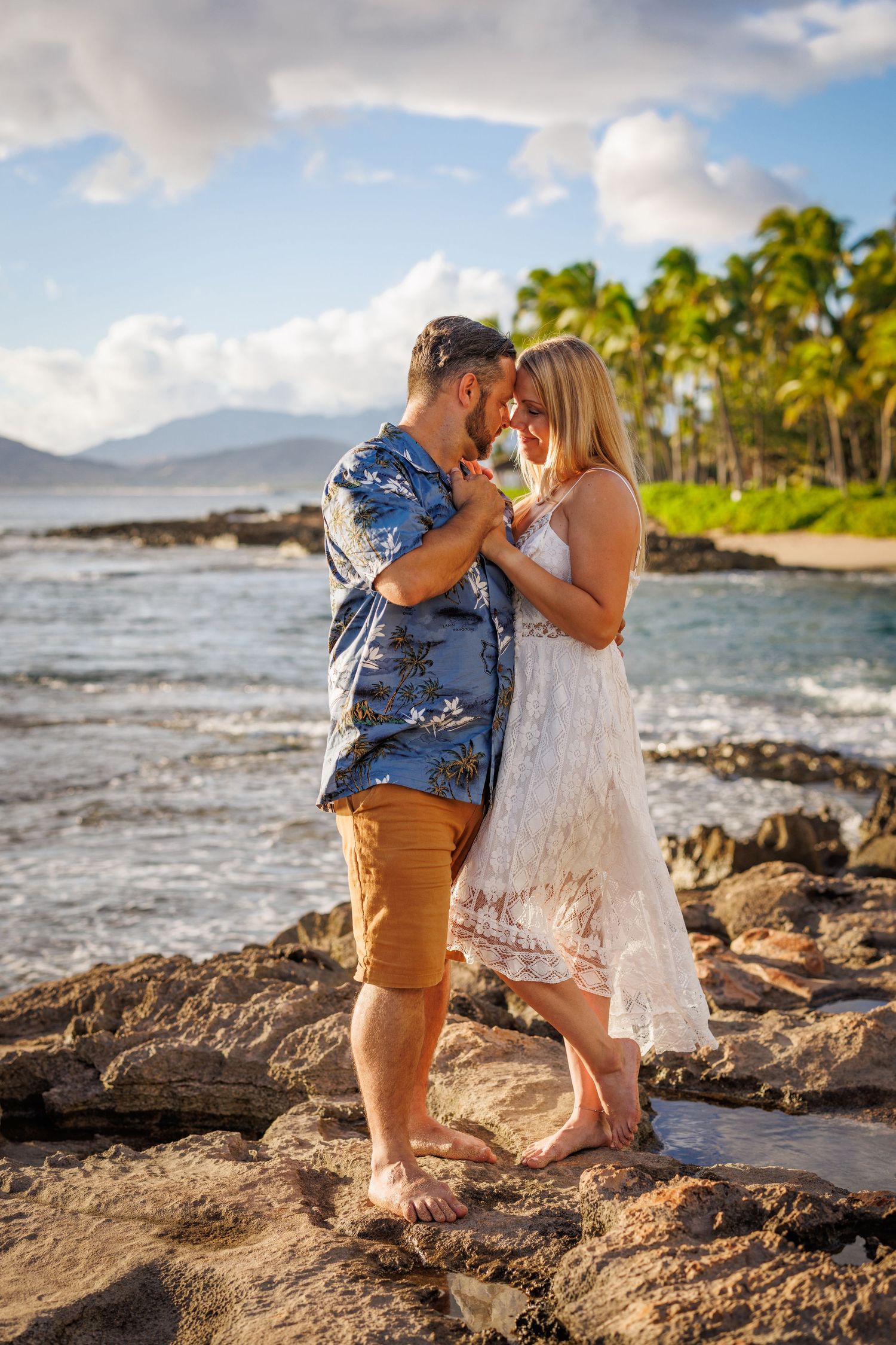 Couple embracing on rocky Hawaiian beach at sunset, palm trees and ocean waves in background.