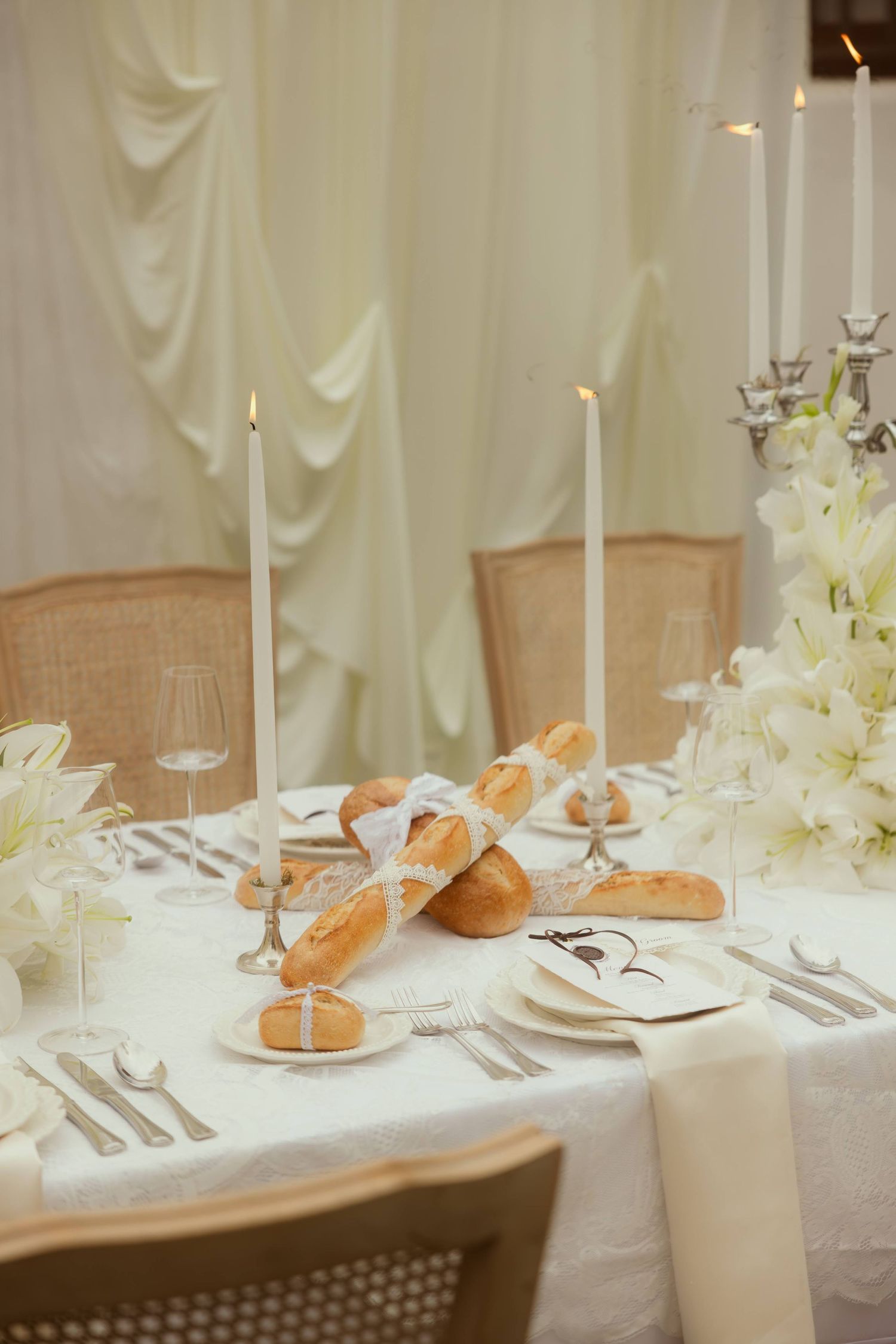 Elegant white wedding table setting with candles, bread rolls, white flowers, and fine silverware on a linen tablecloth.
