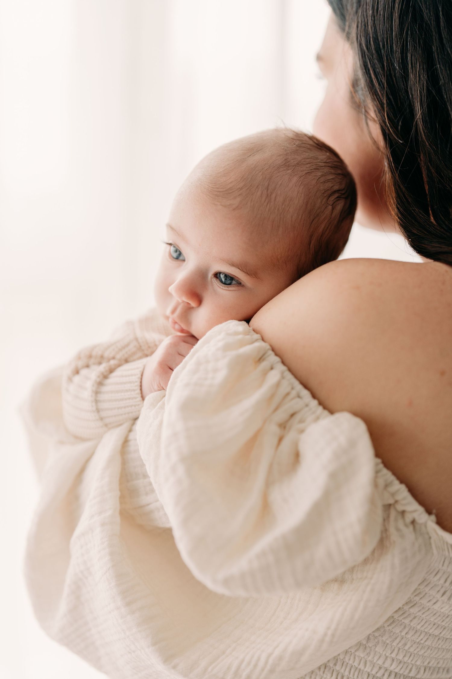 Plan rapproché sur le visage d’un bébé aux yeux bleus dans les bras de sa maman portant une robe de mousseline crème lors d’une séance