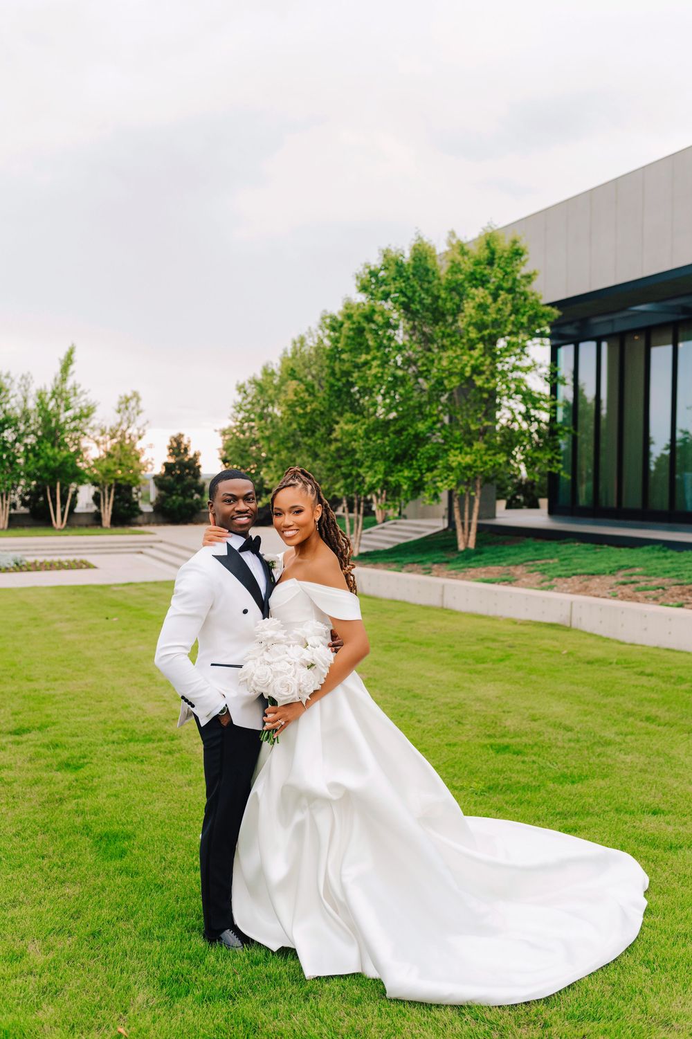 Bride and groom pose on green lawn, she in off-shoulder ball gown, he in white tuxedo jacket.