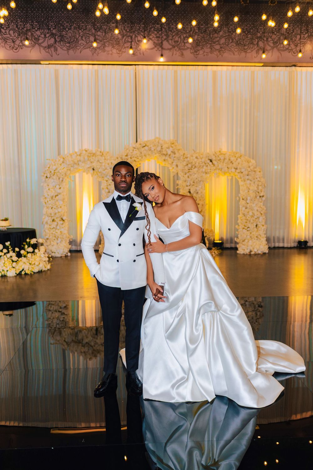 Elegant bride and groom posing at a luxurious wedding reception with floral arch and string lights backdrop.