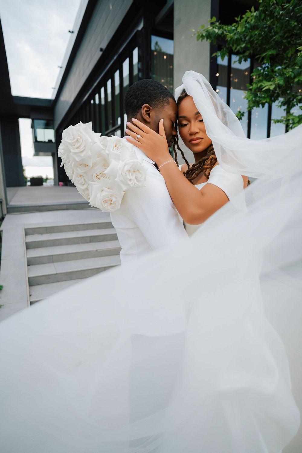 Black couple in wedding attire embrace tenderly, bride in flowing white veil and gown, groom in white floral suit outdoors.
