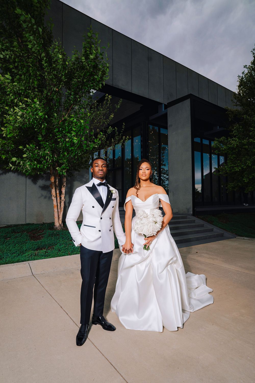 Elegant Black couple in wedding attire pose outside modern building, groom in white tuxedo, bride in off-shoulder satin gown.