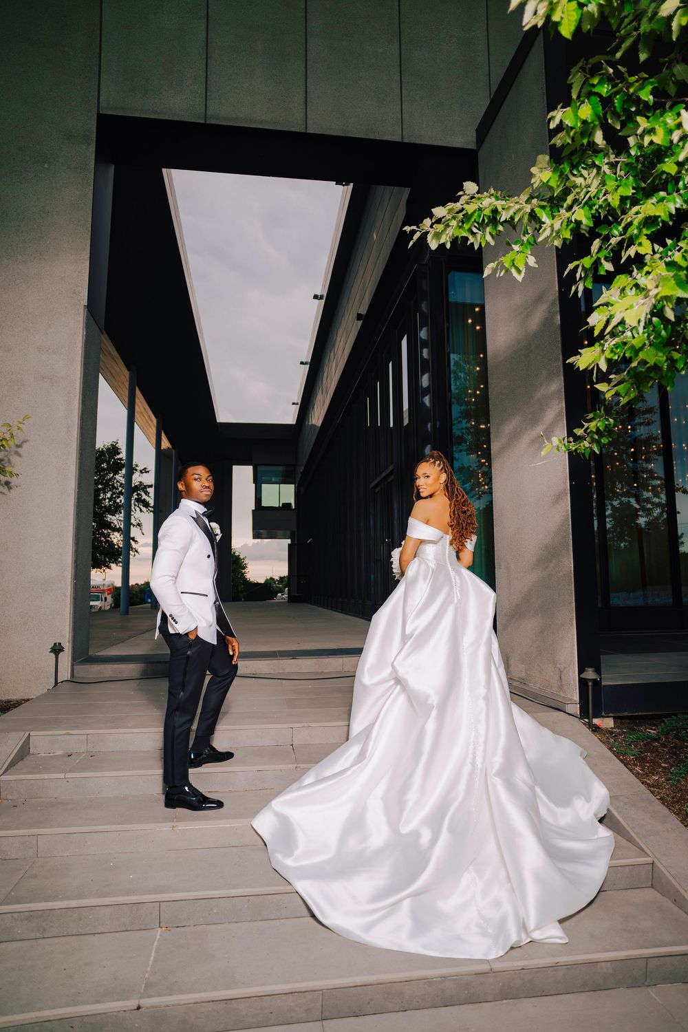 Elegant Black couple in wedding attire pose on modern building steps, bride's satin gown trailing beautifully.