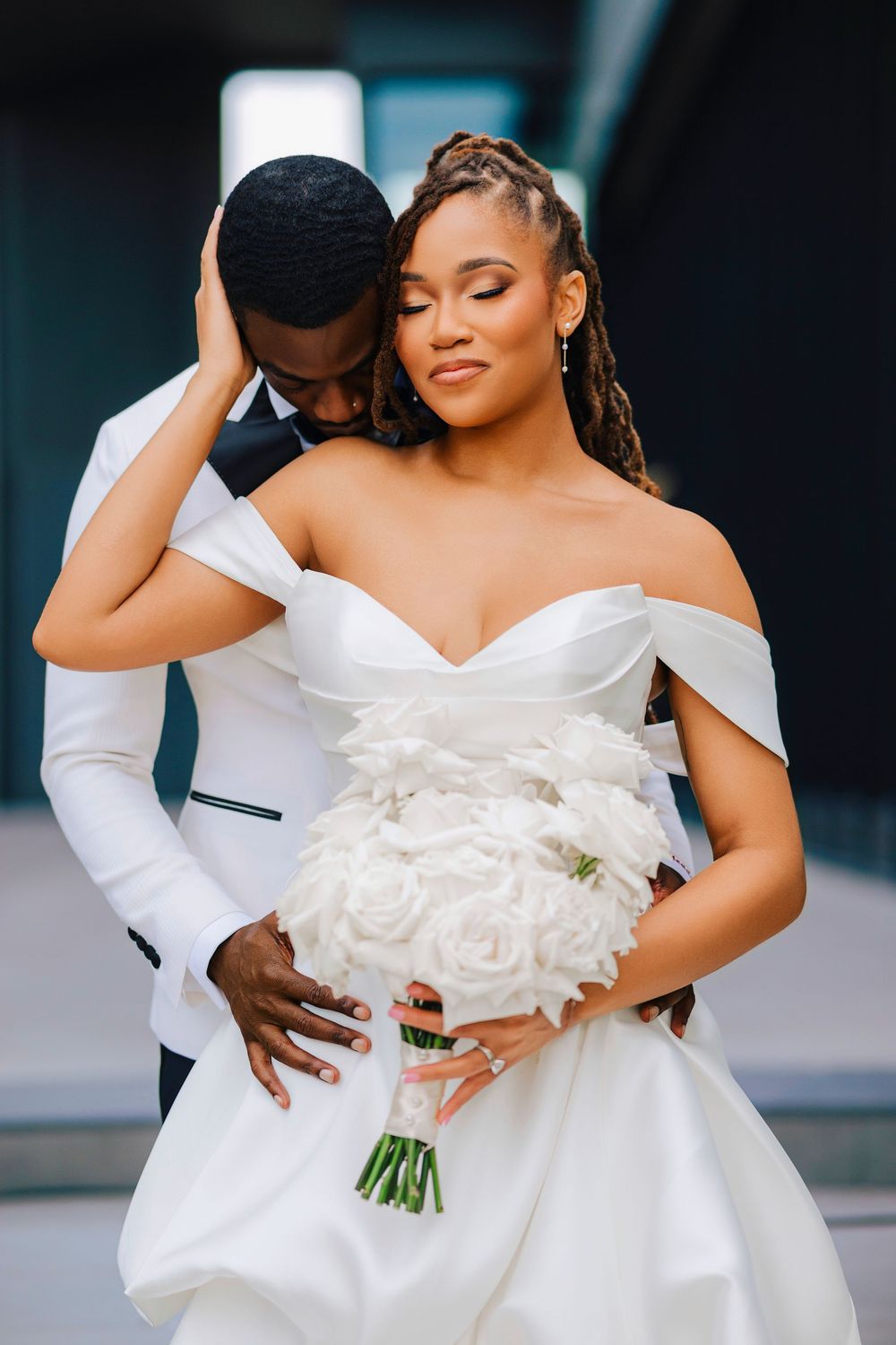 Bride in off-shoulder white gown holds rose bouquet as groom embraces her tenderly.