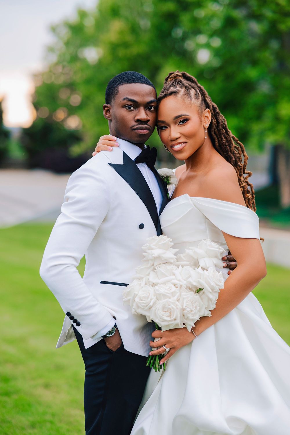 Bride holding white rose bouquet and groom in white tuxedo pose together at outdoor wedding.