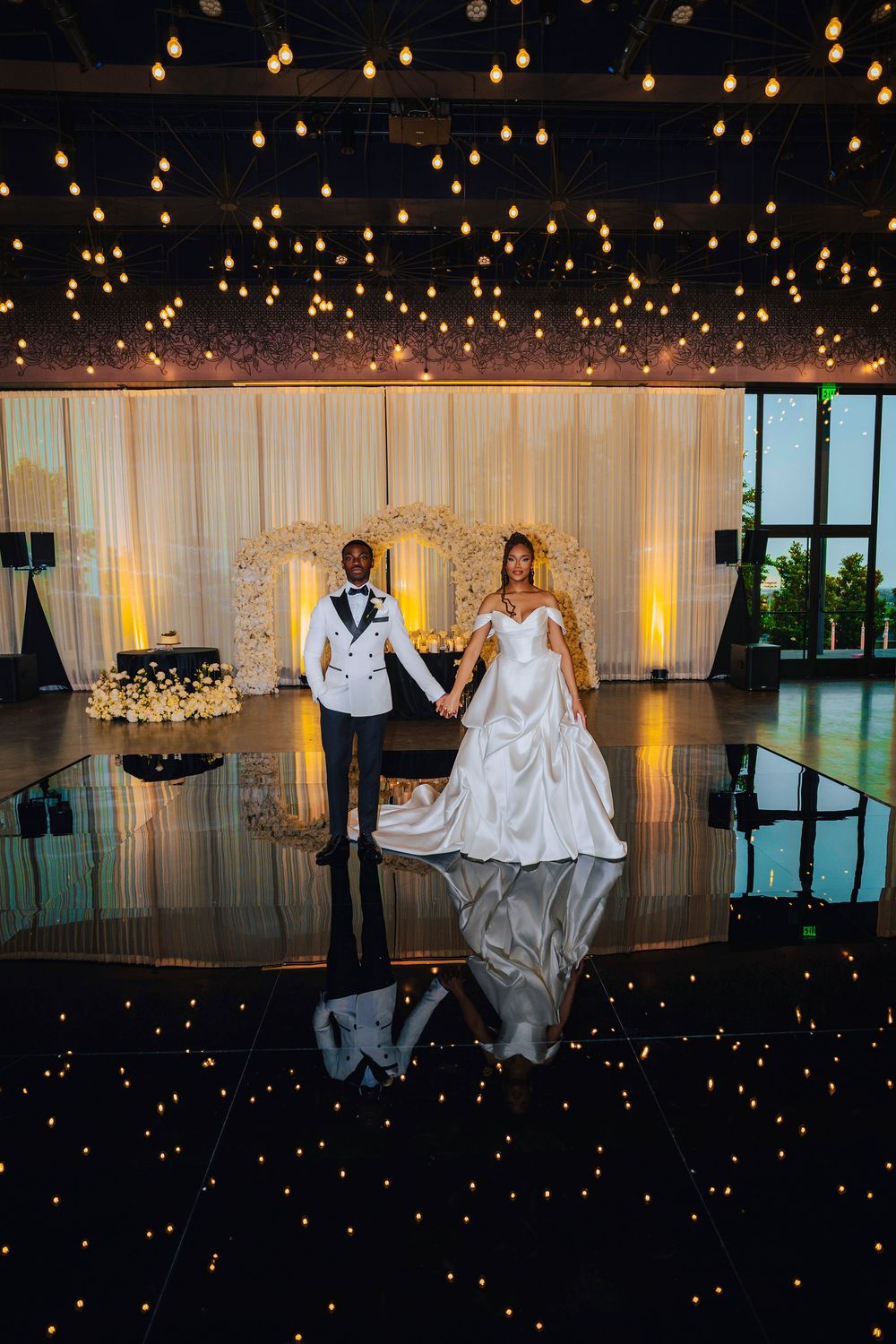 Bride and groom holding hands on a reflective dance floor surrounded by twinkling lights at an elegant wedding reception.