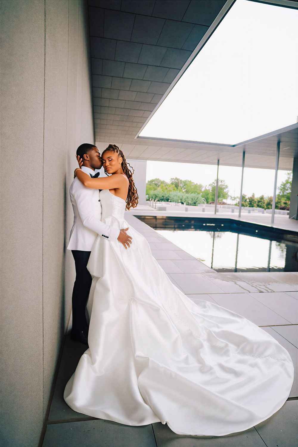 Bride and groom embrace against concrete wall, her dramatic satin train cascading beside a pool.