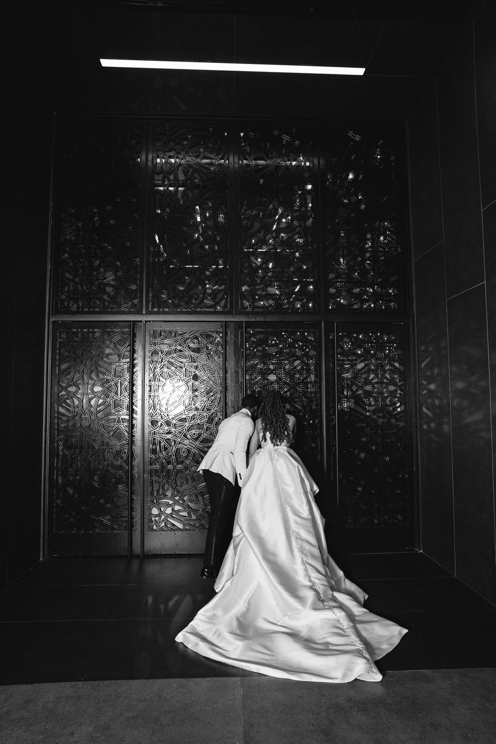 Black and white photo of bride in flowing satin gown walking toward ornate decorative doors.