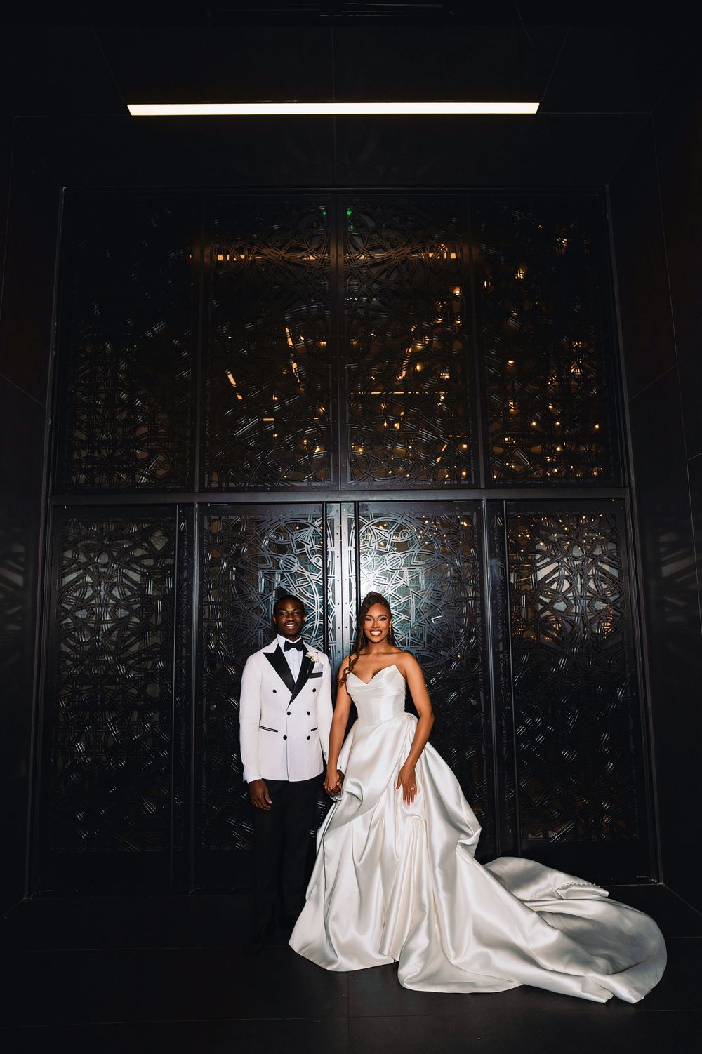 Bride and groom pose before ornate dark decorative doors, groom in white tuxedo jacket.