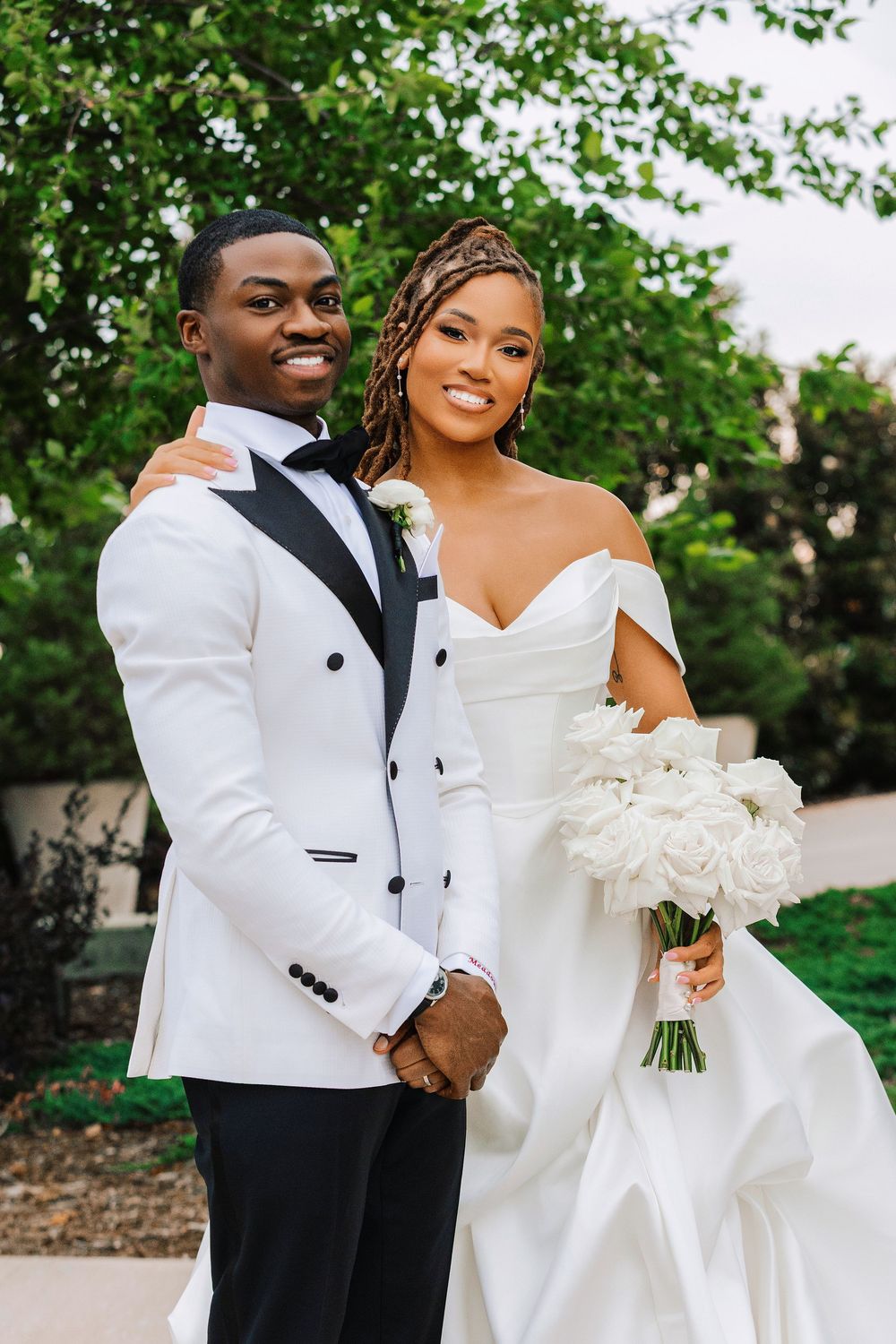 A smiling bride in an off-shoulder white gown holds white roses beside her groom in a white tuxedo outdoors.