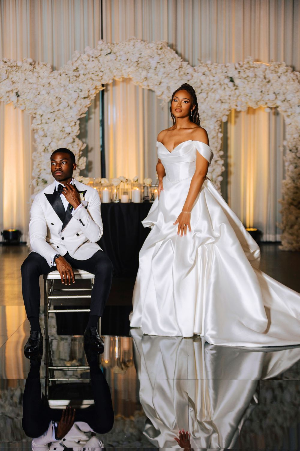 Bride in voluminous white satin gown stands beside seated groom in white tuxedo before romantic floral wedding backdrop.