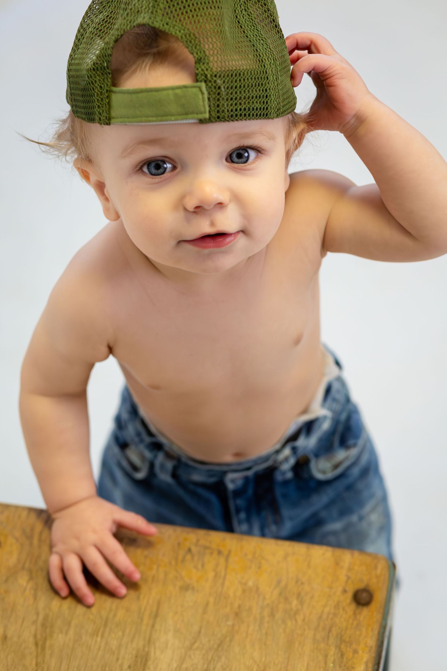Adorable baby in jeans and green trucker cap standing at wooden table against white background.