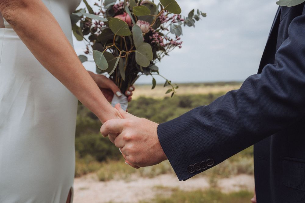 Bride and groom holding hands at outdoor wedding ceremony, bride holding eucalyptus bouquet with pink flowers.