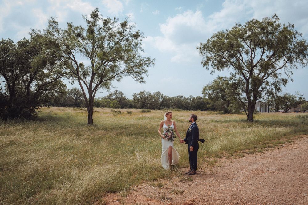 Bride and groom standing together on a rural dirt path surrounded by green trees and open fields.
