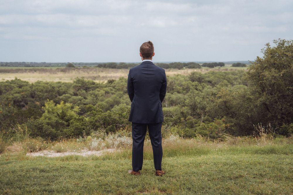 A man in a navy suit stands with his back to the camera, overlooking a vast green landscape under a cloudy sky.