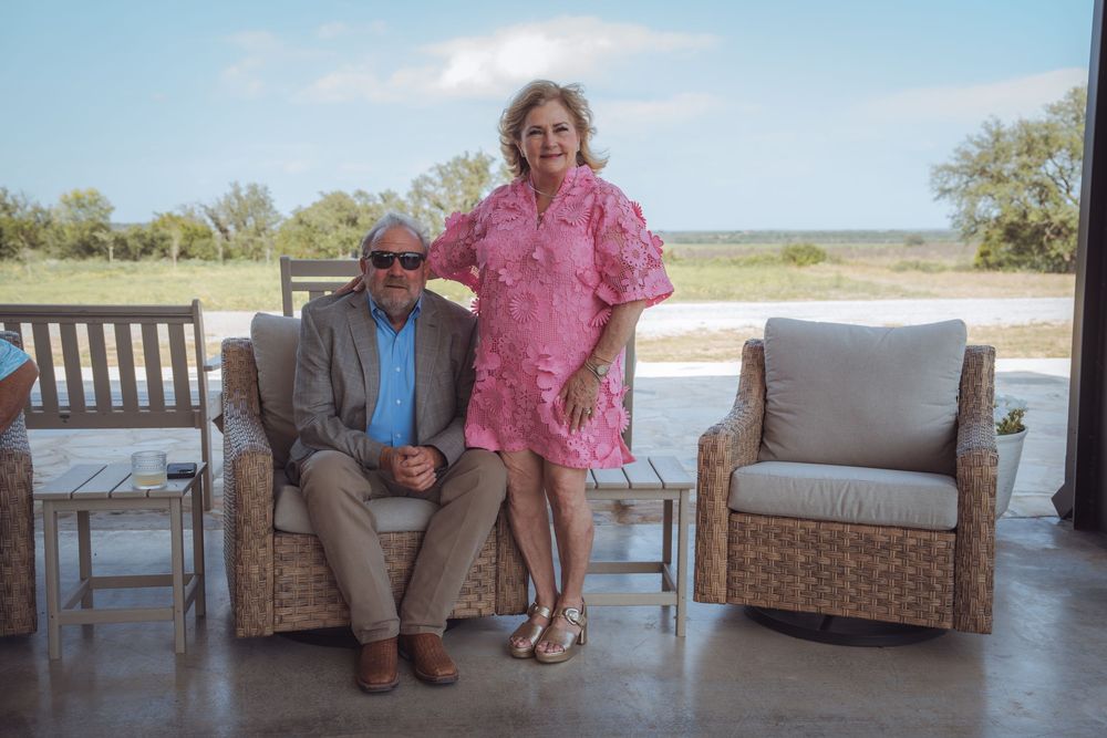Couple posing on a covered patio with wicker furniture, man in blazer seated, woman in pink dress standing, rural landscape behind.
