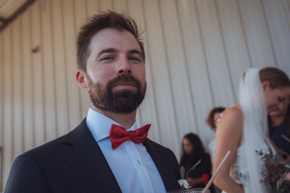 Bearded groom in navy suit and red bow tie smiling at wedding ceremony.