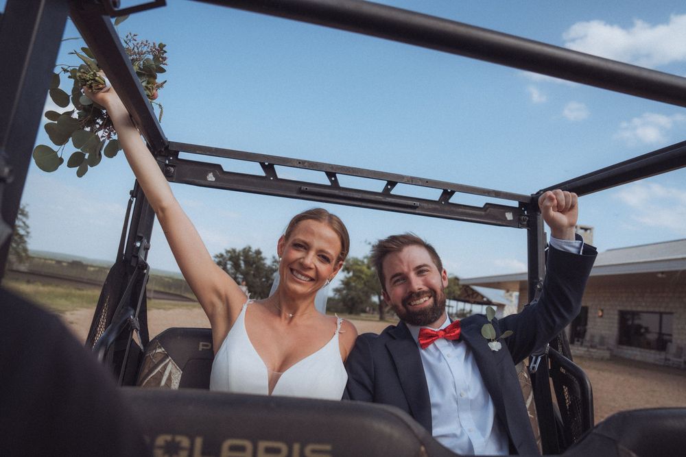 Happy newlyweds celebrate in an open-top jeep, bride in white dress and groom in navy suit with red bow tie.